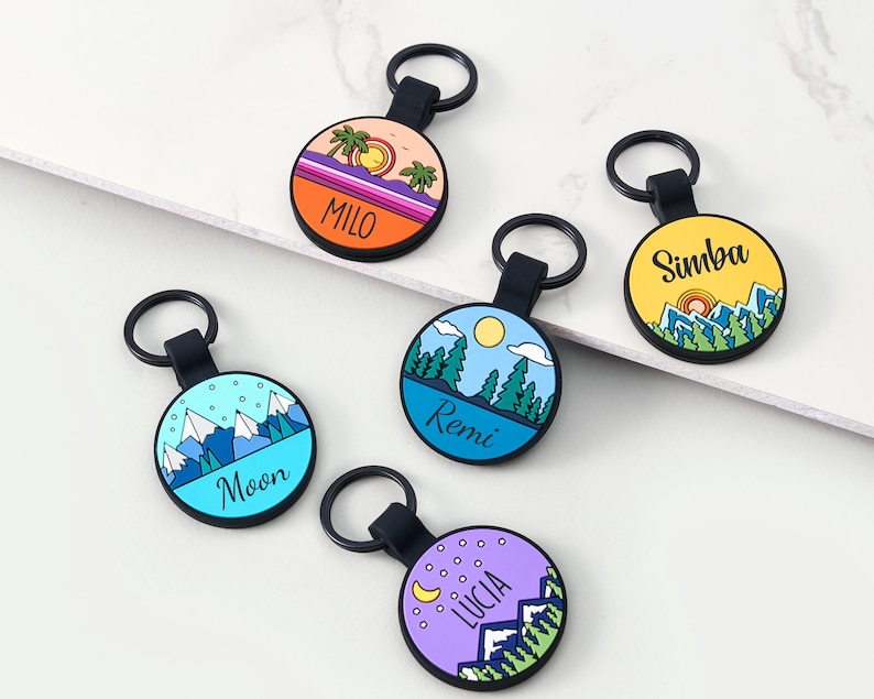 May include: A selection of personalised keyrings with black straps and key rings. Each keyring has a colourful circular design with a name and a landscape illustration, including mountains, trees, and a sunset. The names on the keyrings are Milo, Simba, Remi, Moon, and Lucia.