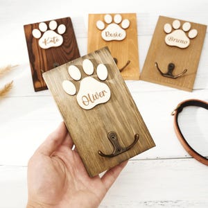 Puede incluir: Cuatro ganchos de pared rectangulares de madera con un recorte de huella de pata de madera clara en la parte superior. Cada huella está grabada con un nombre: Kate, Rosie, Bruno y Oliver. Cada placa de madera incluye un gancho doble de metal bronce.
