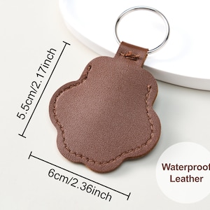 May include: A brown, paw-shaped leather keychain with a silver ring. The keychain is 2.36 inches wide and 2.17 inches tall. The text "Waterproof Leather" is visible. The keychain is designed for pet lovers.