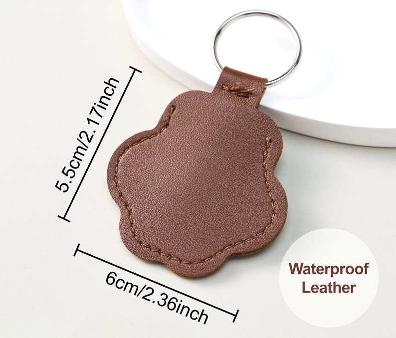 May include: A brown, paw-shaped leather keychain with a silver ring. The keychain is 2.36 inches wide and 2.17 inches tall. The text "Waterproof Leather" is visible. The keychain is designed for pet lovers.