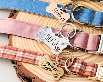 May include: Close-up of three dog collars with personalized bone-shaped tags. The collars are denim blue, pink velvet, and red plaid. Tags are engraved with names and paw prints. The tags are attached with metal rings.