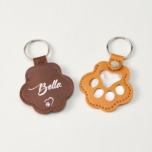 May include: Two paw-shaped keychains. One is brown with the name "Bella" in script and a heart with a paw print. The other is tan with a clear insert. Both have silver key rings.