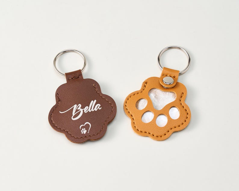 May include: Two paw-shaped keychains. One is brown with the name "Bella" in script and a heart with a paw print. The other is tan with a clear insert. Both have silver key rings.