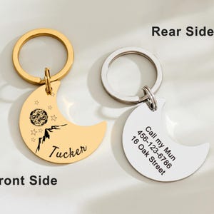 May include: Two crescent moon-shaped pet ID tags with key rings. One is gold-colored with a mountain and star design, the name "Tucker", and a planet graphic. The other is silver-colored with contact information.