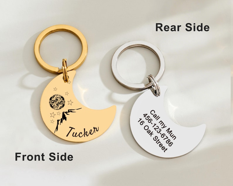 May include: Two crescent moon-shaped pet ID tags with key rings. One is gold-colored with a mountain and star design, the name "Tucker", and a planet graphic. The other is silver-colored with contact information.