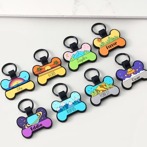 May include: A collection of colorful, bone-shaped pet ID tags with black key rings. Each tag features a unique design, including landscapes, sunsets, and space themes, with personalized names like "Sky" and "Eddie" in bold text.