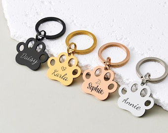 May include: A collection of personalized pet ID tags in the shape of a paw print. The tags are in black, gold, rose gold, and silver, each with a different name engraved. Each tag has a matching keyring.