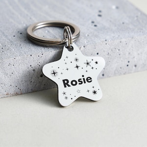 Puede incluir: Un llavero blanco en forma de estrella con el nombre "Rosie" en letras negras. La estrella est&aacute; decorada con dise&ntilde;os de estrellas y brillos negros. El llavero tiene un anillo de metal plateado.
