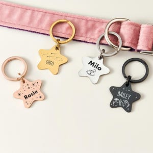 May include: Four star-shaped pet tags with names and designs. The tags are gold, rose gold, silver, and black, each attached to a keyring. The tags are personalized with the names "Rosie", "Oreo", "Milo", and "Bailey". A pink dog collar is in the background.