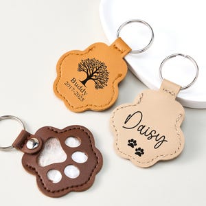 May include: Three leather paw-shaped keychains. One is tan with a tree graphic and the name "Buddy" and dates. Another is light tan with the name "Daisy" and paw prints. The third is brown with a paw print cutout.
