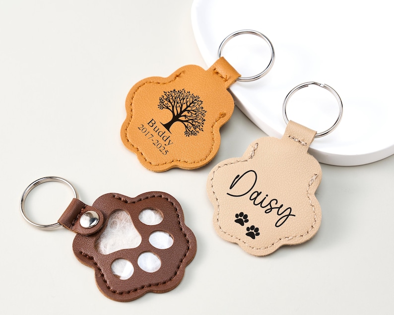 May include: Three leather paw-shaped keychains. One is tan with a tree graphic and the name "Buddy" and dates. Another is light tan with the name "Daisy" and paw prints. The third is brown with a paw print cutout.
