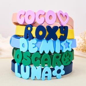 May include: A stack of five colorful silicone collars, each featuring raised lettering and a symbol. The collars are pink with COCO and a heart, yellow with ROXY, light blue with DEMI and a star, green with OSCAR and a paw print, and navy with LUNA and a flower.