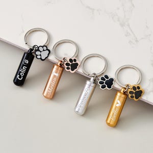 May include: Four metal pet urn keychains in black, rose gold, silver, and gold. Each keychain has a small cylindrical urn with a name engraved on it and a paw print charm. The keychains are on a white and gray surface.