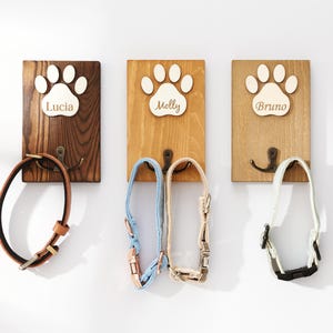 Puede incluir: Tres ganchos de pared rectangulares de madera con acabados en marrón oscuro, medio y claro. Cada uno presenta una huella de pata de madera blanca con los nombres grabados Lucia, Molly y Bruno. Un gancho de metal color bronce sostiene collares.