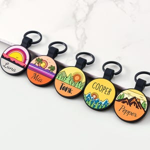 May include: Five circular keyrings with black loops, each displaying a landscape design and a name. Designs include a sunset, palm trees, mountains, and forests, with names such as "Luna" and "Mia".