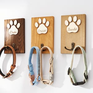 Può includere: Tre ganci da parete rettangolari in legno con intagli a forma di zampa, ciascuno dotato di un gancio in metallo e un'etichetta con i nomi Duke, Oscar o Scarlet. I ganci sostengono un collare in pelle marrone, uno blu, uno beige e uno verde chiaro.
