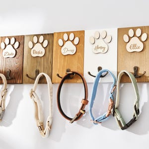Puede incluir: Un soporte de pared para correas de mascotas hecho de madera con cinco paneles rectangulares en varios tonos de madera y blanco. Cada panel muestra una huella de pata de madera grabada con los nombres Duke, Rosie, Daisy, Honey y Ellie, con ganchos.