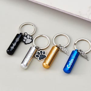 May include: Four keyrings with cylindrical pendants. One black, one silver, one gold, and one blue. Each keyring has a silver ring and a charm. The black keyring has a paw print charm and the name "Archie". The silver keyring has a paw print charm and the name "Zoe". The gold keyring has wing charms and the name "Coco". The blue keyring has wing charms and the name "Colin".