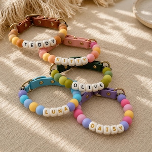 May include: Five pet collars featuring colorful matte beads and white letter beads spelling out the names TEDDY, KITTY, OLIVE, LUNA, and DAISY. Each collar includes a leather-like strap with a metal buckle and gold-toned hardware accents.