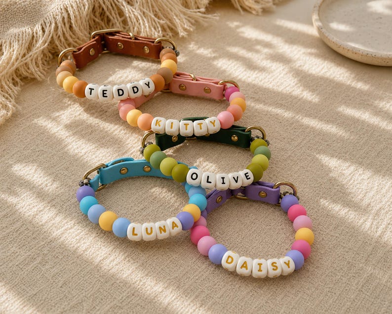 May include: Five pet collars featuring colorful matte beads and white letter beads spelling out the names TEDDY, KITTY, OLIVE, LUNA, and DAISY. Each collar includes a leather-like strap with a metal buckle and gold-toned hardware accents.