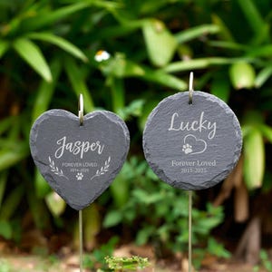 May include: Two slate pet memorial garden stakes. One is heart-shaped, the other round. The heart-shaped stake reads "Jasper Forever Loved 2014-2025". The round stake reads "Lucky Forever Loved 2015-2025" with a paw print.