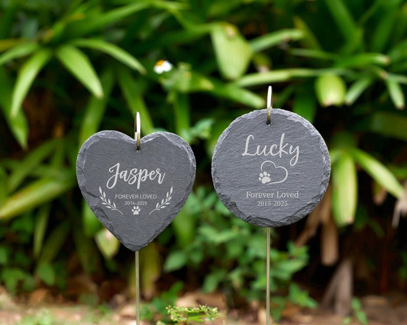 May include: Two slate pet memorial garden stakes. One is heart-shaped, the other round. The heart-shaped stake reads "Jasper Forever Loved 2014-2025". The round stake reads "Lucky Forever Loved 2015-2025" with a paw print.