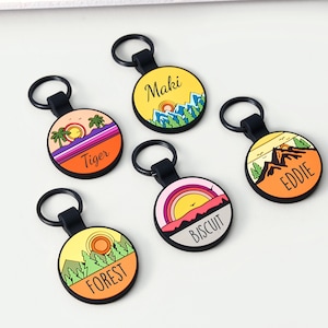 May include: Five circular keyrings with black loops. Each keyring displays a colourful landscape design with text: "Maki", "Tiger", "Eddie", "Biscuit", and "Forest". The designs include mountains, sunsets, and trees.
