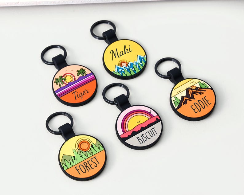 May include: Five circular keyrings with black loops. Each keyring displays a colourful landscape design with text: "Maki", "Tiger", "Eddie", "Biscuit", and "Forest". The designs include mountains, sunsets, and trees.