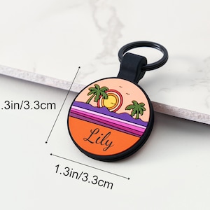 May include: A circular keychain with a beach scene design. The keychain is 3.3 cm in diameter and features a sunset with palm trees, a sun, and the name "Lily" in black lettering. The keychain has a black ring.