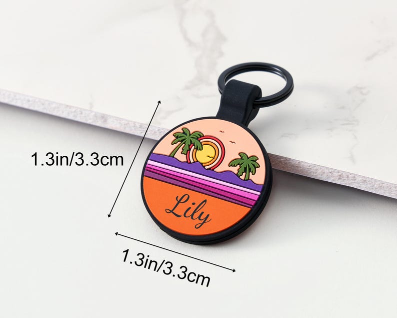 May include: A circular keychain with a beach scene design. The keychain is 3.3 cm in diameter and features a sunset with palm trees, a sun, and the name "Lily" in black lettering. The keychain has a black ring.