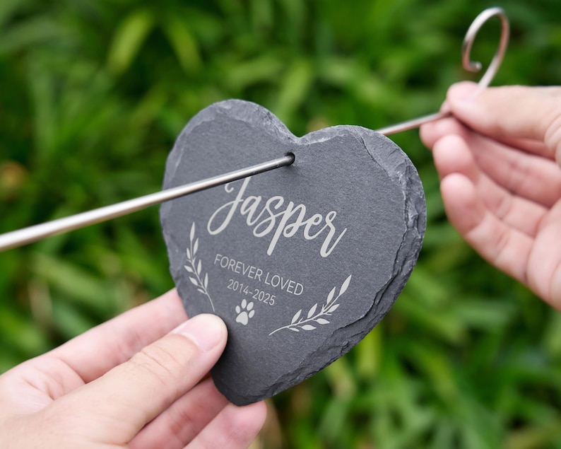 May include: A heart-shaped, dark gray slate pet memorial stake with the name "Jasper" in elegant script. The text "FOREVER LOVED 2014-2025" and a paw print are also engraved. A metal stake is attached for garden placement.