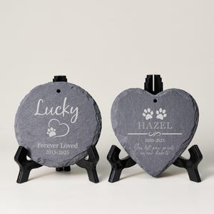 May include: Two slate pet memorial stones. One is round with the name "Lucky", a paw print, and "Forever Loved 2015-2025". The other is heart-shaped with "Hazel", paw prints, and "2020-2025". Both are on black stands.