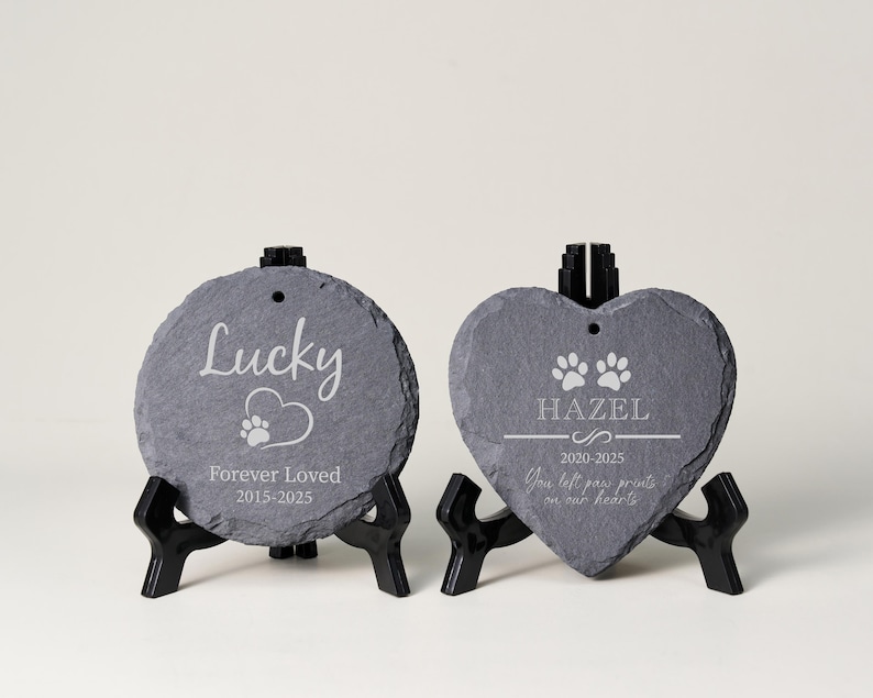 May include: Two slate pet memorial stones. One is round with the name "Lucky", a paw print, and "Forever Loved 2015-2025". The other is heart-shaped with "Hazel", paw prints, and "2020-2025". Both are on black stands.