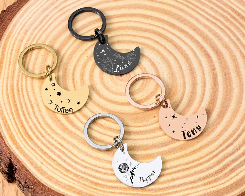 May include: Five crescent moon keychains in gold, black, silver, and rose gold. Each keychain has a ring and is engraved with a name and star or floral designs. The names are "Toffee", "Luna", "Tony", and "Pepper".