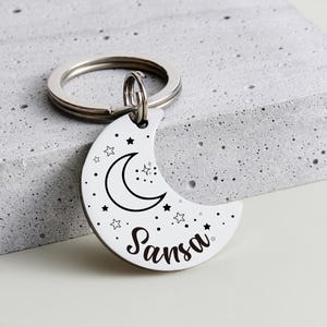 May include: A white crescent moon keychain with a black outline of a moon, stars, and the name "Sansa" in a script font. The keychain has a silver ring and is resting on a gray surface.