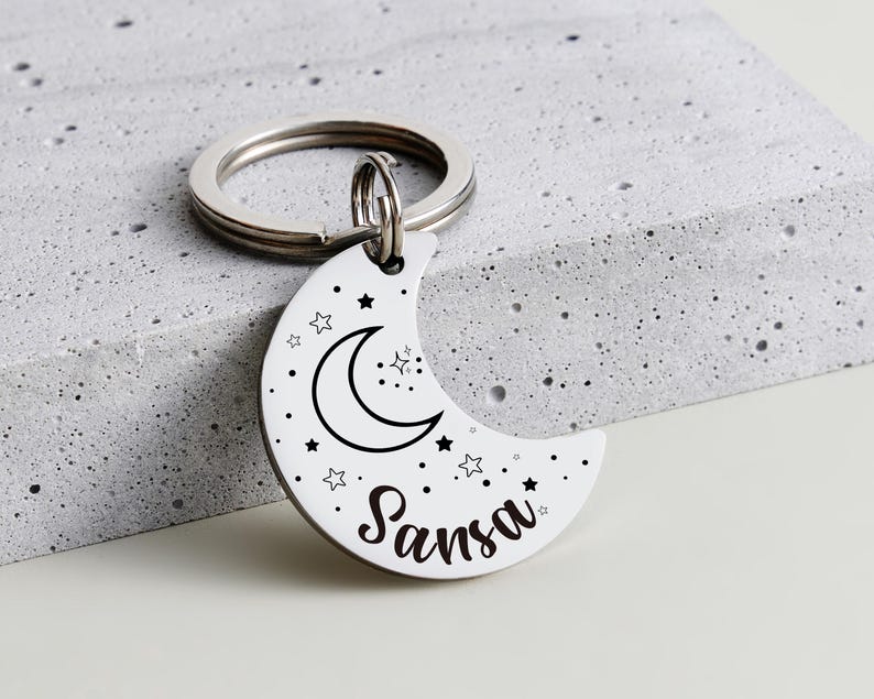 May include: A white crescent moon keychain with a black outline of a moon, stars, and the name "Sansa" in a script font. The keychain has a silver ring and is resting on a gray surface.