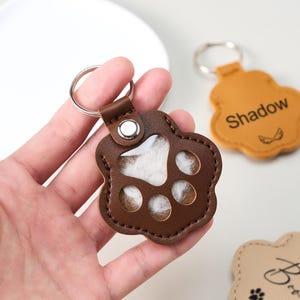 May include: A brown leather paw-shaped keychain with a silver keyring. The keychain has a clear window revealing white fur inside. Another keychain in the background is tan with the name "Shadow" and small wing graphics.