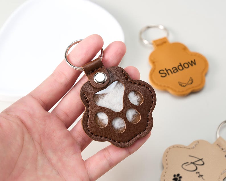 May include: A brown leather paw-shaped keychain with a silver keyring. The keychain has a clear window revealing white fur inside. Another keychain in the background is tan with the name "Shadow" and small wing graphics.