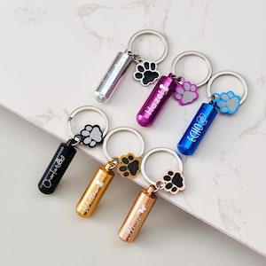 Peut inclure: Plusieurs porte-clés commémoratifs pour animaux de compagnie, de couleurs variées : argent, violet, bleu, noir, or et or rose. Chaque porte-clés est orné d'un charm en forme de patte et d'un petit cylindre gravé d'un nom.