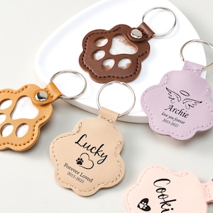 May include: A collection of paw-shaped keychains in various colors, including brown, tan, and pink. Some keychains feature a paw print cutout, while others have personalized text such as "Lucky" and "Archie." Each keychain has a metal ring for attaching keys.