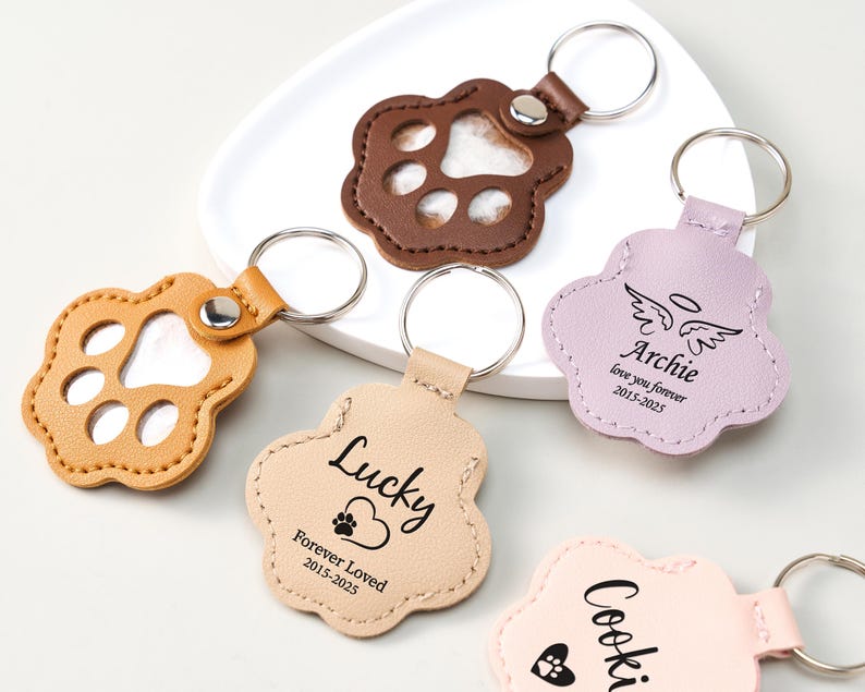 May include: A collection of paw-shaped keychains in various colors, including brown, tan, and pink. Some keychains feature a paw print cutout, while others have personalized text such as "Lucky" and "Archie." Each keychain has a metal ring for attaching keys.