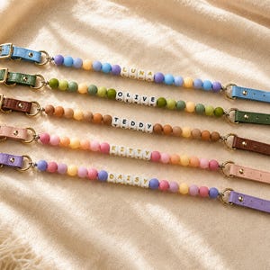 May include: Five beaded pacifier clips featuring colorful silicone beads and white letter cubes spelling the names LUNA, OLIVE, TEDDY, KITTY, and DAISY. Each clip has a gold-tone metal buckle and a leather-like strap in blue, green, brown, pink, or purple.