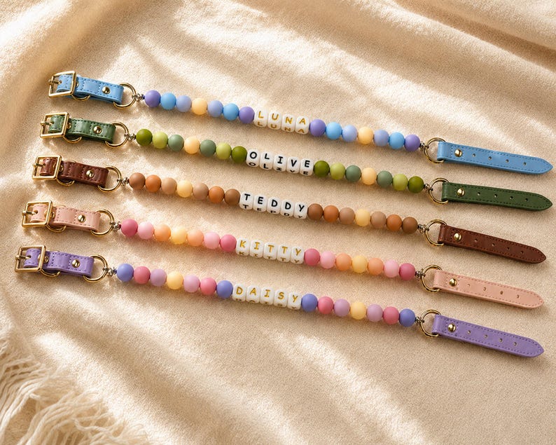May include: Five beaded pacifier clips featuring colorful silicone beads and white letter cubes spelling the names LUNA, OLIVE, TEDDY, KITTY, and DAISY. Each clip has a gold-tone metal buckle and a leather-like strap in blue, green, brown, pink, or purple.