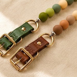 May include: Two lanyards featuring matte silicone beads in shades of green, yellow, and brown. Each lanyard includes a gold-toned metal buckle, a matching ring, and a leather-like strap with a gold-toned rivet, providing a secure attachment point.