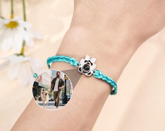May include: A turquoise braided bracelet with a silver charm in the shape of a flower. The charm has a black center. A small circular image of a person running with a dog is visible.