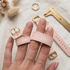 May include: Two light pink leather straps with gold-toned metal buckles. One strap is labeled with a width of 1.5 cm and the other with a width of 2.0 cm. A gold-toned metal caliper tool is positioned nearby for scale, alongside a small gold ring.