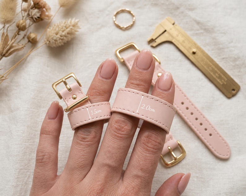 May include: Two light pink leather straps with gold-toned metal buckles. One strap is labeled with a width of 1.5 cm and the other with a width of 2.0 cm. A gold-toned metal caliper tool is positioned nearby for scale, alongside a small gold ring.