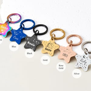 Puede incluir: Una colecci&oacute;n de etiquetas de identificaci&oacute;n para mascotas en forma de estrella, en varios colores: arco&iacute;ris, azul, negro, dorado, oro rosa y plateado. Cada etiqueta presenta el nombre de una mascota y un dise&ntilde;o &uacute;nico, con un llavero.