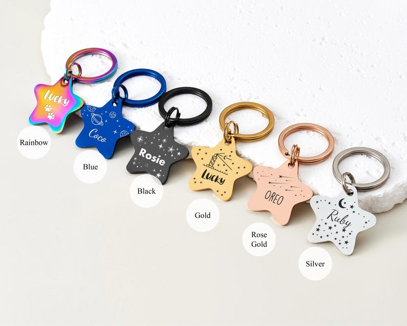 Puede incluir: Una colecci&oacute;n de etiquetas de identificaci&oacute;n para mascotas en forma de estrella, en varios colores: arco&iacute;ris, azul, negro, dorado, oro rosa y plateado. Cada etiqueta presenta el nombre de una mascota y un dise&ntilde;o &uacute;nico, con un llavero.
