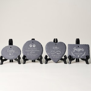 May include: Four slate pet memorial stones in oval, round, heart, and rectangular shapes, each on a black stand. The stones are engraved with pet names, dates, paw prints, and memorial text. The text is in a white color.